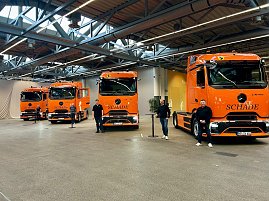 "Vier orangefarbene Mercedes-Benz eActros-Lkw mit Schade-Logo nebeneinander in einer Halle aufgereiht, Personen stehen davor.