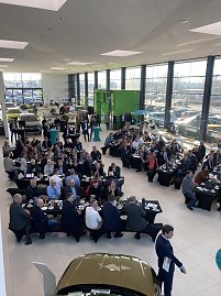 Weitwinkelaufnahme der Skoda Autowelt Peter mit vielen Sitzgruppen, an denen G&auml;ste fr&uuml;hst&uuml;cken. Rechts und links stehen Autos, hinten gro&szlig;e Fensterfront mit Tageslicht.