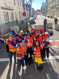 Gruppe von als Narren verkleideten Menschen in orangefarbenen Warnwesten steht auf einer Stra&szlig;e und winkt fr&ouml;hlich in die Kamera.