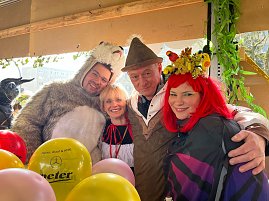 "Vier Personen in farbenfrohen Kost&uuml;men � darunter ein B&auml;renkost&uuml;m, ein Dirndl und ein Fantasiekost&uuml;m mit roter Per&uuml;cke � stehen beieinander und l&auml;cheln in die Kamera, umgeben von bunten Luftballons.