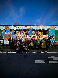 Gro&szlig;e Gruppe verkleideter Menschen posiert vor und auf einem bunt gestalteten Karnevalswagen mit M&auml;rchenmotiven, viele halten bunte Luftballons.