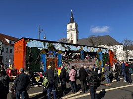 Karnevalswagen mit Burg- und Wolkenmotiven f&auml;hrt durch eine Innenstadt, Menschen stehen am Stra&szlig;enrand und schauen zu, im Hintergrund eine Kirche mit Turm.