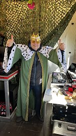 "Mann in gr&uuml;ner K&ouml;nigskutte mit Hermelinbesatz und goldener Krone steht lachend vor einem Mischpult und hebt beide Daumen hoch.