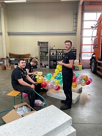 Drei M&auml;nner in schwarzer Arbeitskleidung sitzen und stehen in einer Werkstatt, binden bunte Autohaus-Peter-Luftballons zusammen, im Hintergrund ein orangefarbener LKW.