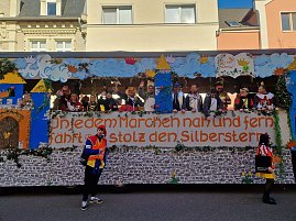 Seitenansicht des bunt gestalteten Karnevalswagens mit dem Schriftzug �In jedem M&auml;rchen nah und fern f&auml;hrt er stolz den Silberstern�, mehrere verkleidete Menschen stehen darauf, zwei Personen in Narrenkost&uuml;men davor.
