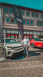 Ein Mann steht neben einem wei&szlig;en Citro&euml;n-Ausstellungswagen vor einem Peugeot-Banner, im Hintergrund ein roter Opel.
