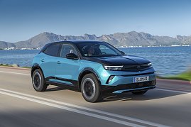 Türkis-schwarzer Opel Mokka Electric fährt an einer Küstenstraße entlang, Meer und Gebirge im Hintergrund, Blick schräg von vorn links.