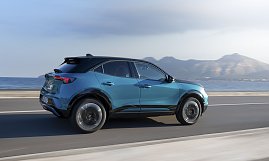 Türkis-schwarzer Opel Mokka Electric fährt an einer Küstenstraße entlang, Meer und Gebirge im Hintergrund, Blick schräg von hinten links.