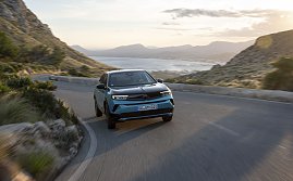 Türkis-schwarzer Opel Mokka Electric fährt auf einer kurvigen Küstenstraße bergauf, Blick frontal von vorn, Meer und Berge im Hintergrund.