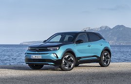 Türkis-schwarzer Opel Mokka Electric steht am Meer, Blick schräg von vorn links, Berge im Hintergrund.