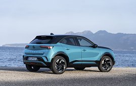 Türkis-schwarzer Opel Mokka Electric steht am Meer, Blick schräg von hinten links, Berge im Hintergrund.