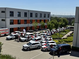 Gesamtansicht des DRK-Fuhrparks: Luftaufnahme des Innenhofs vor einem DRK-Geb&auml;ude mit zahlreichen Einsatzfahrzeugen, einem Rettungswagen und Mitarbeitenden, die neben den Autos stehen.