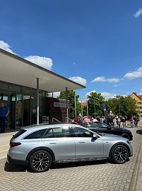 Vor der Stadthalle Wittenberg steht ein silberner Mercedes-Benz Kombi. Im Hintergrund sind weitere Autos, Menschen und eine Mercedes-Fahne zu sehen.