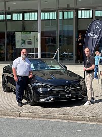 Zwei M&auml;nner stehen vor einem schwarzen Mercedes-Benz Coup&eacute; und l&auml;cheln in die Kamera. Rechts im Hintergrund ist eine Mercedes-Fahne, dahinter Glasfassade der Stadthalle.