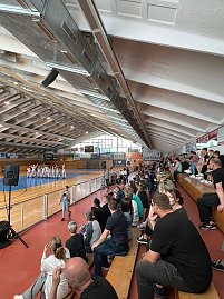 Innenaufnahme einer Sporthalle mit gef&uuml;llter Trib&uuml;ne. Auf der Spielfl&auml;che stehen in Formation mehrere Sportlerinnen in wei&szlig;en Anz&uuml;gen bei einer Vorf&uuml;hrung.