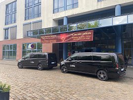 Shuttlebusse vor Hotel: Zwei schwarze Mercedes-Benz Shuttlebusse mit Autohaus Peter-Logo stehen vor dem Radisson Blu Fürst Leopold Hotel in Dessau, vor einem Banner zur Veranstaltung Music meets Dinner.