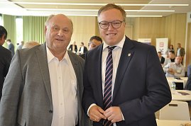 Das Bild zeigt links Geschäftsführer Helmut Peter und rechts Mario Voigt. Beide stehen nebeneinander in einem Konferenzraum, lächeln in die Kamera und tragen Anzüge. Im Hintergrund sind weitere Personen an Tischen zu sehen.