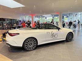 Ein weißes Mercedes-Cabriolet mit der Aufschrift She’s Mercedes steht in einem Autohaus, im Hintergrund sind Gäste und Personal in roten T-Shirts zu sehen.