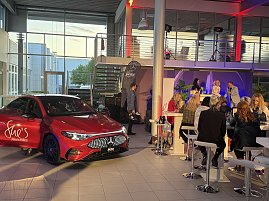 Roter Mercedes-Benz CLA 250+ steht in einer Veranstaltungshalle, im Hintergrund sitzen Gäste an Stehtischen. Mercedes-Benz CLA 250+ mit EQ-Technologie | Energieverbrauch kombiniert: 14,1-12,2 kWh/100 km | CO2Emissionen kombiniert: 0 g/km | CO2-Emissionsklasse: A