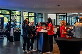 Mehrere Frauen, teils in roten She’s Mercedes-T-Shirts, teils in schwarzen Outfits, stehen an einem Stehtisch mit Gläsern und unterhalten sich in einem hellen Autohaus.