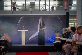 Christina Dost spricht auf einer Bühne mit Mercedes-Logo im Hintergrund, rotem Teppich davor und Publikum auf beiden Seiten.