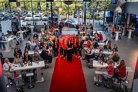 Eine große Gruppe von Frauen sitzt und steht um Tische in einem Autohaus, in der Mitte posiert das She’s Mercedes-Team auf dem roten Teppich.