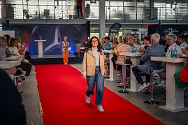 Eine junge Frau mit dunklen, langen Haaren, Brille, beiger Jacke, hellblauem Oberteil, blauen Jeans und weißen Sneakern läuft bei einer Modenschau über einen roten Teppich.