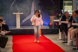 Eine junge Frau mit langen, dunklen Haaren, rosa Bluse, hellen Jeans und beiger Handtasche läuft lächelnd über den roten Teppich.