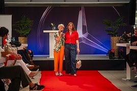 Moderatorin in farbenfrohem Blazer mit einer Frau im roten Oberteil, gestreiftem Shirt und weiter Jeans am Ende des Laufstegs.