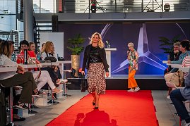 Frau mit blonden Haaren, schwarzem Oberteil, schwarzer Jacke und Leopardenrock trägt eine schwarze Handtasche über den Laufsteg.