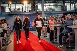 Mehrere Models in sportlicher Kleidung – schwarze Jacken mit roten und weißen Akzenten – laufen gleichzeitig über den roten Teppich, ein junger Mann und eine junge Frau vorne, weitere Personen dahinter.