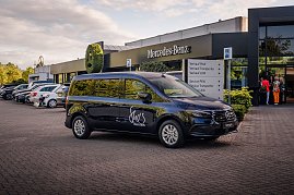 Schwarzer Mercedes-Van mit She’s Mercedes-Logo vor einem Mercedes-Benz Autohaus geparkt. Im Hintergrund stehen weitere Fahrzeuge und einige Menschen am Eingang.