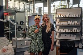 Zwei Frauen lächeln in die Kamera; die linke trägt ein gemustertes Kleid und eine gelbe Kappe, die rechte einen schwarzen Anzug. Im Hintergrund Regale mit Kosmetikprodukten.