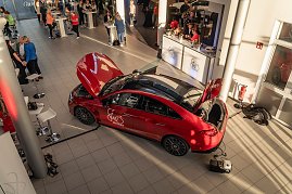 Von oben fotografierter roter Mercedes mit She’s Mercedes-Logo, Motorhaube und Kofferraum geöffnet. Im Autohaus sind Besucher an Stehtischen und einem Catering-Stand zu sehen. Mercedes-Benz CLA 250+ mit EQ-Technologie | Energieverbrauch kombiniert: 14,1-12,2 kWh/100 km | CO2Emissionen kombiniert: 0 g/km | CO2-Emissionsklasse: A
