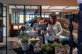 Zwei Frauen betrachten und riechen Pflanzen an einem Floristik-Stand im Autohaus, eine hält ein Glas mit rosafarbenem Getränk.