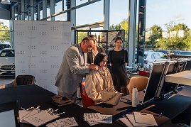Friseurmeister in grauem Anzug hält die Haare einer sitzenden Frau und schlägt ihr eine Frisur vor, während zwei weitere Frauen zusehen. Vor ihnen stehen Kosmetik- und Styling-Utensilien auf Tischen.