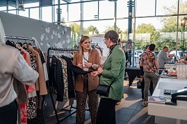 Zwei Frauen unterhalten sich an einem Kleiderständer mit verschiedenen Jacken und Blusen. Die Frau links trägt einen braunen Blazer, die Frau rechts einen grünen Blazer.