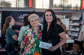 Geschäftsführerin Christina Dost im schwarzen Hosenanzug und Moderatorin Mandy Kossira im bunten, geblümten Blazer lächeln gemeinsam in die Kamera, Christina Dost hält eine Karte mit She’s Mercedes-Logo.