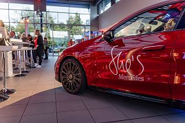 Seitliche Nahaufnahme eines roten Mercedes mit She’s Mercedes-Logo, im Hintergrund Menschen an Stehtischen im Autohaus. Mercedes-Benz CLA 250+ mit EQ-Technologie | Energieverbrauch kombiniert: 14,1-12,2 kWh/100 km | CO2Emissionen kombiniert: 0 g/km | CO2-Emissionsklasse: A