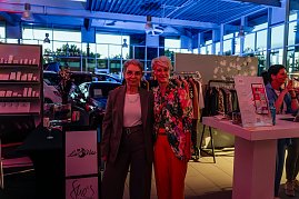 Zwei Frauen stehen im Autohaus vor einem Kleiderständer und lächeln in die Kamera, links eine in grauem Hosenanzug, rechts Mandy Kossira in bunter Jacke mit Blumenmuster.