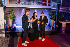 Drei Personen stehen auf rotem Teppich auf einer Bühne, Christina Dost spricht ins Mikrofon, während ein Mann ein Los aus einem goldenen Hut zieht.