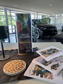 Dekoration zum S&uuml;dafrikanischen Abend im Autohaus Peter: Ein Aufsteller mit der Aufschrift �Herzlich Willkommen zum S&uuml;dafrikanischen Abend� und s&uuml;dafrikanischer Flagge, daneben Flyer mit Safari-Motiven und ein Untersetzer mit Leopardenmuster. Im Hintergrund steht ein schwarzer Mercedes im Showroom.