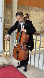 Ein Musiker in einem schwarzen Frack spielt konzentriert auf einem Cello. Die Szene spielt im Innenbereich eines historischen Gebäudes mit Steinwänden und einem rotbraunen Teppich. Der Musiker steht am Geländer einer Galerie, während er mit dem Bogen über die Saiten des Instruments streicht.