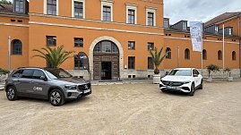 Zwei Mercedes-Benz SUVs, ein graues und ein weißes Modell, parken vor einem historischen Gebäude mit orangefarbener Fassade. Vor dem Eingang stehen Palmen in großen Pflanzkübeln und ein Werbefähnchen von Autohaus Peter.