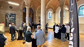 Innenaufnahme eines historischen Saals mit hohen Steinsäulen und Holzboden. Gäste stehen an Stehtischen, unterhalten sich und genießen Getränke. Im Hintergrund ist ein Buffet aufgebaut.