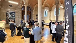 Innenaufnahme eines historischen Saals mit hohen Steinsäulen und Holzboden. Gäste stehen an Stehtischen, unterhalten sich und genießen Getränke. Im Hintergrund ist ein Buffet aufgebaut.