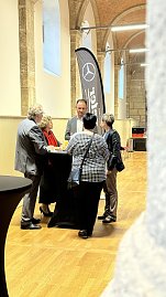 Eine kleine Gruppe von fünf Personen unterhält sich im historischen Saal an einem schwarzen Stehtisch. Im Hintergrund sind hohe Steinsäulen, Wände aus Naturstein und ein großes Mercedes-Benz Werbefähnchen zu sehen.