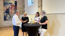 Vier Personen stehen und unterhalten sich an einem schwarzen Stehtisch in einem historischen Saal mit Holzboden und Steinwänden. Auf dem Tisch stehen Gläser mit Sekt und Orangensaft. Im Hintergrund hängt ein farbenfrohes Gemälde an der Wand.