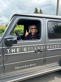 Eine lächelnde Frau mit Sonnenbrille sitzt am Steuer eines grauen Mercedes-Benz G-Klasse Geländewagens, lehnt sich aus dem offenen Fenster und zeigt den Daumen hoch. Auf der Fahrzeugseite ist die Aufschrift THE GELÄNDEWAGEN und Mercedes-Benz gut sichtbar.