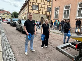 Zwei Mitarbeiter der Peter Gruppe stehen lächelnd auf der Straße während des Festumzugs in Sondershausen. Hinter ihnen fahren Fahrzeuge der Firma, am Straßenrand stehen Zuschauer.